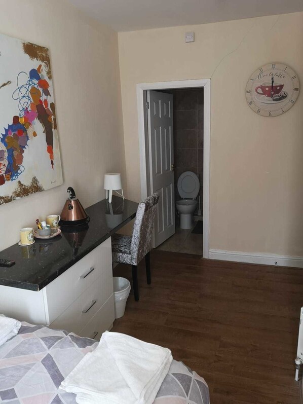 A bedroom with a bed, a desk with a kettle, a chair, a painting, and a clock on the wall.