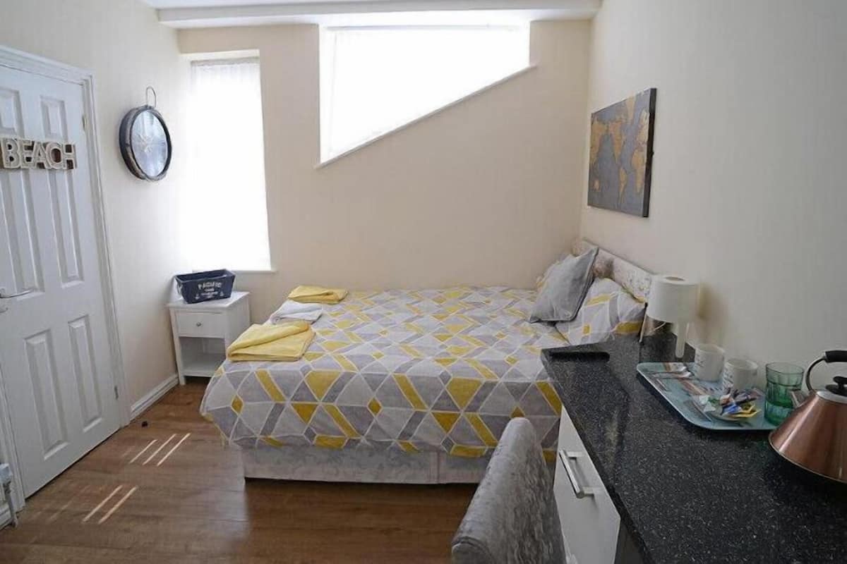 Une chambre avec un lit, une table de chevet, une fenêtre, un tableau et un coin cuisine.