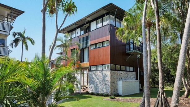 Un immeuble moderne à plusieurs étages, doté d’une façade en pierre, de balcons et d’un jardin bien entretenu avec des palmiers.