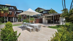 Une terrasse extérieure moderne, dotée d’un ensemble de mobilier de jardin et d’un espace barbecue, le tout entouré d’une végétation luxuriante et de bâtiments contemporains.