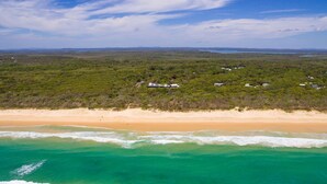 Vue aérienne d’une zone côtière avec une plage de sable et des eaux turquoise. On aperçoit plusieurs bâtiments dispersés dans un paysage verdoyant, qui pourraient appartenir à un complexe hôtelier ou à une propriété de vacances.