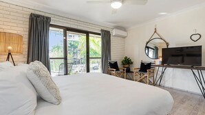 A hotel room with a large bed, a TV, a desk with a chair, and a balcony with a view of greenery.