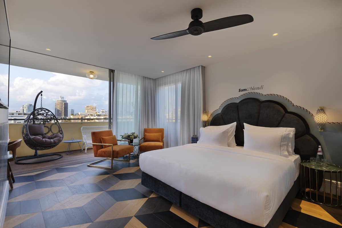 A modern hotel room with a large bed, a ceiling fan, a city view, and a balcony with seating.