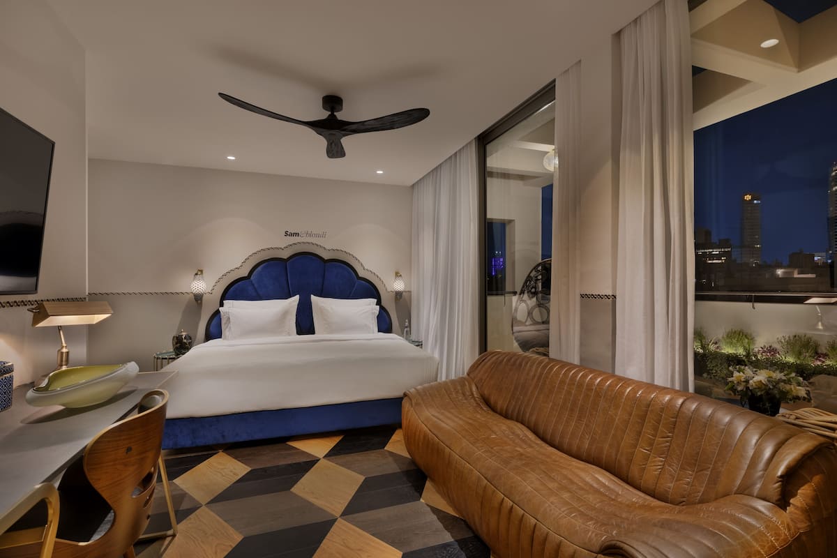 A hotel room with a bed, a leather sofa, a desk with a lamp, and a city view through a large window.