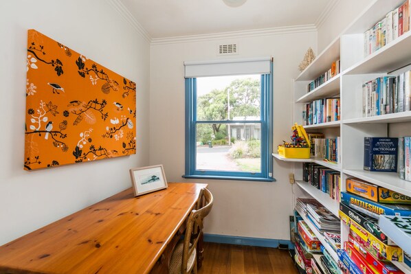 A room with a wooden table, a bookshelf filled with books and games, a window with a view of the outside, and a wall with an orange artwork.