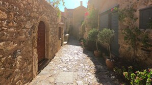 Une étroite ruelle pavée de pierres, bordée de bâtiments rustiques en pierre, dotés d’arcades et agrémentée de plantes en pot.