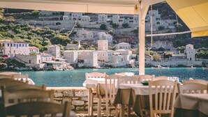 Un village côtier aux bâtiments en pierre, bordé d’une mer d’un bleu éclatant, et doté d’un espace de restauration avec des tables et des chaises.
