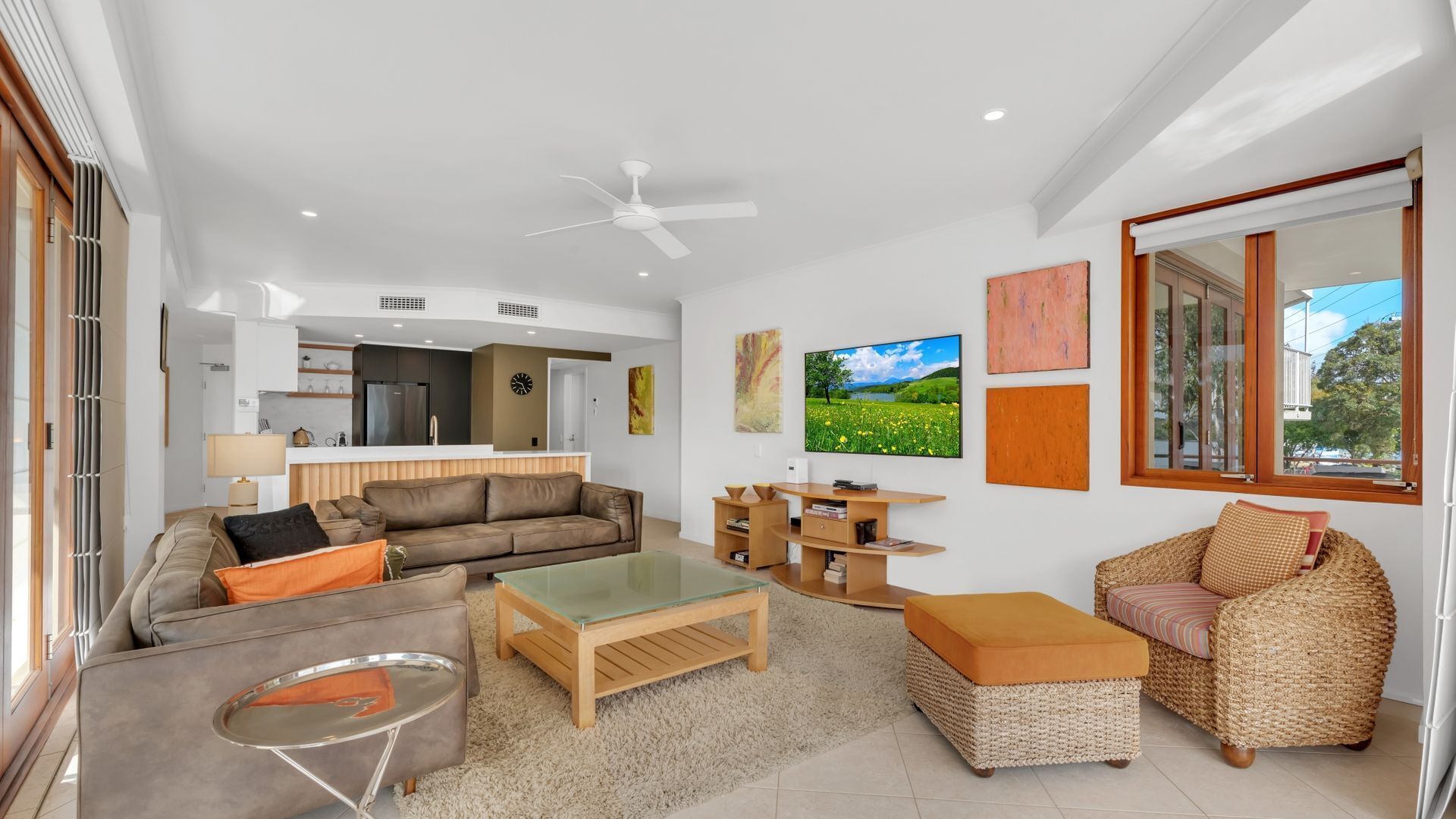 A modern living room with a sofa, coffee table, and wicker chair. There is a kitchen area in the background with a refrigerator and cabinets.