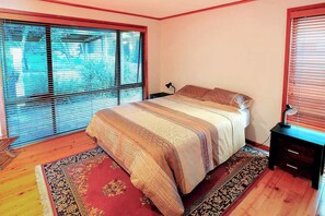 Un dormitorio con una cama, una mesita de noche, una lámpara, una ventana con persianas y una alfombra estampada.