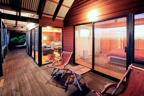 A wooden deck with two lounge chairs and a table, leading to a glass door entrance.