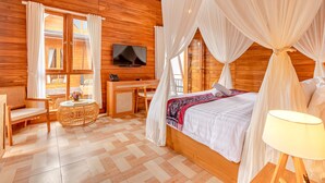 A wooden cabin room with a bed, a desk, a chair, a TV, and a window with curtains. - Kubu GWK Resort (Jimbaran)