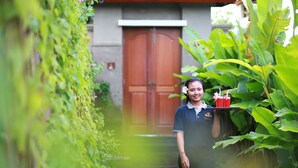 Una persona sostiene una bandeja con bebidas, de pie en un exuberante jardín con una puerta de madera y grandes plantas verdes al fondo.
