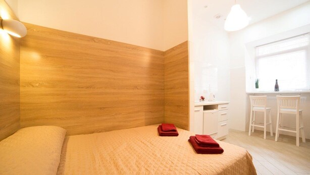 A modern bedroom with a wooden headboard, a bed with beige bedding, and two red towels on the bed. There is a small desk with a chair and a window with blinds. - Smart Apartment Filatova 10a (Lviv)