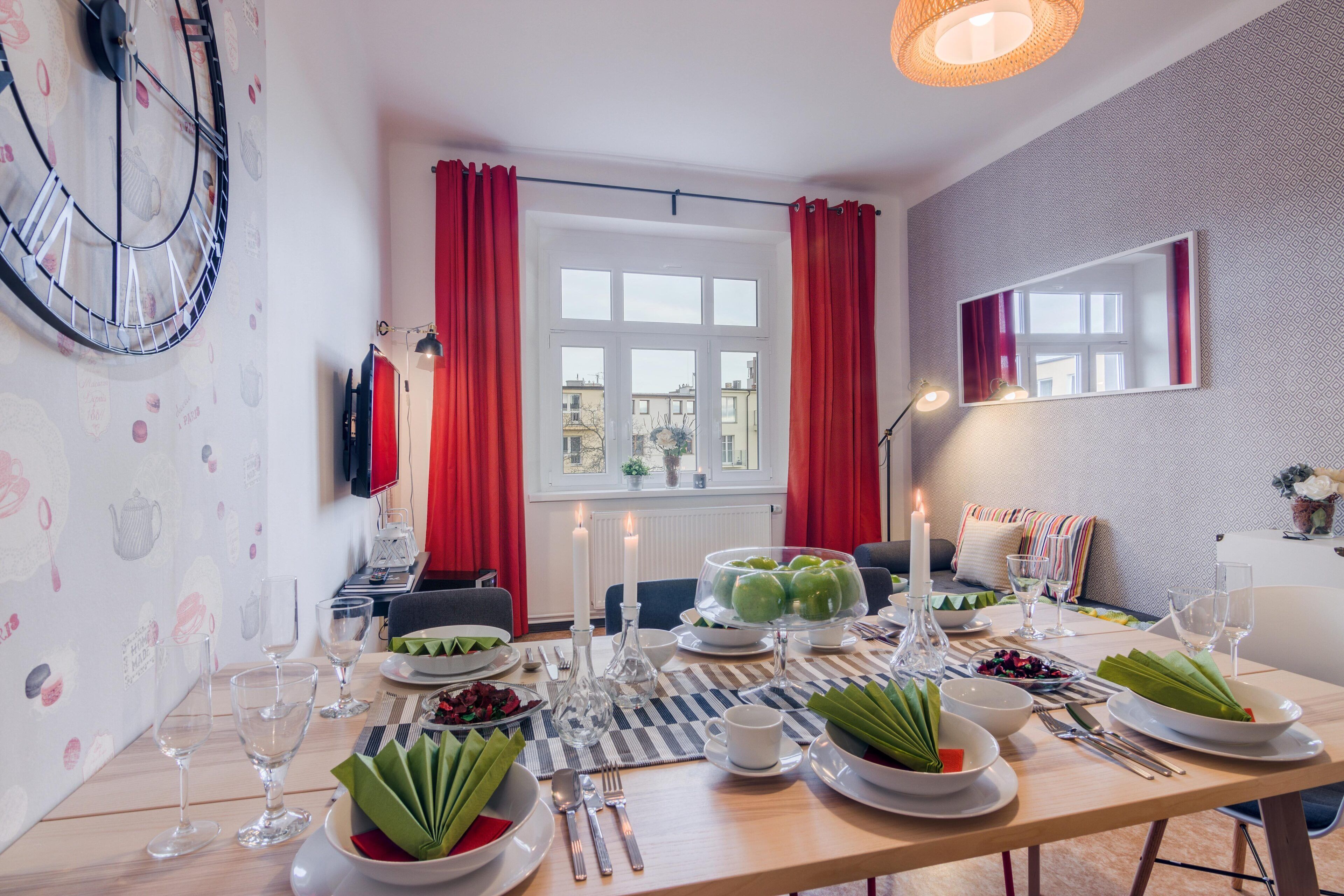 A dining area with a table set for a meal, a clock on the wall, and a window with red curtains.
