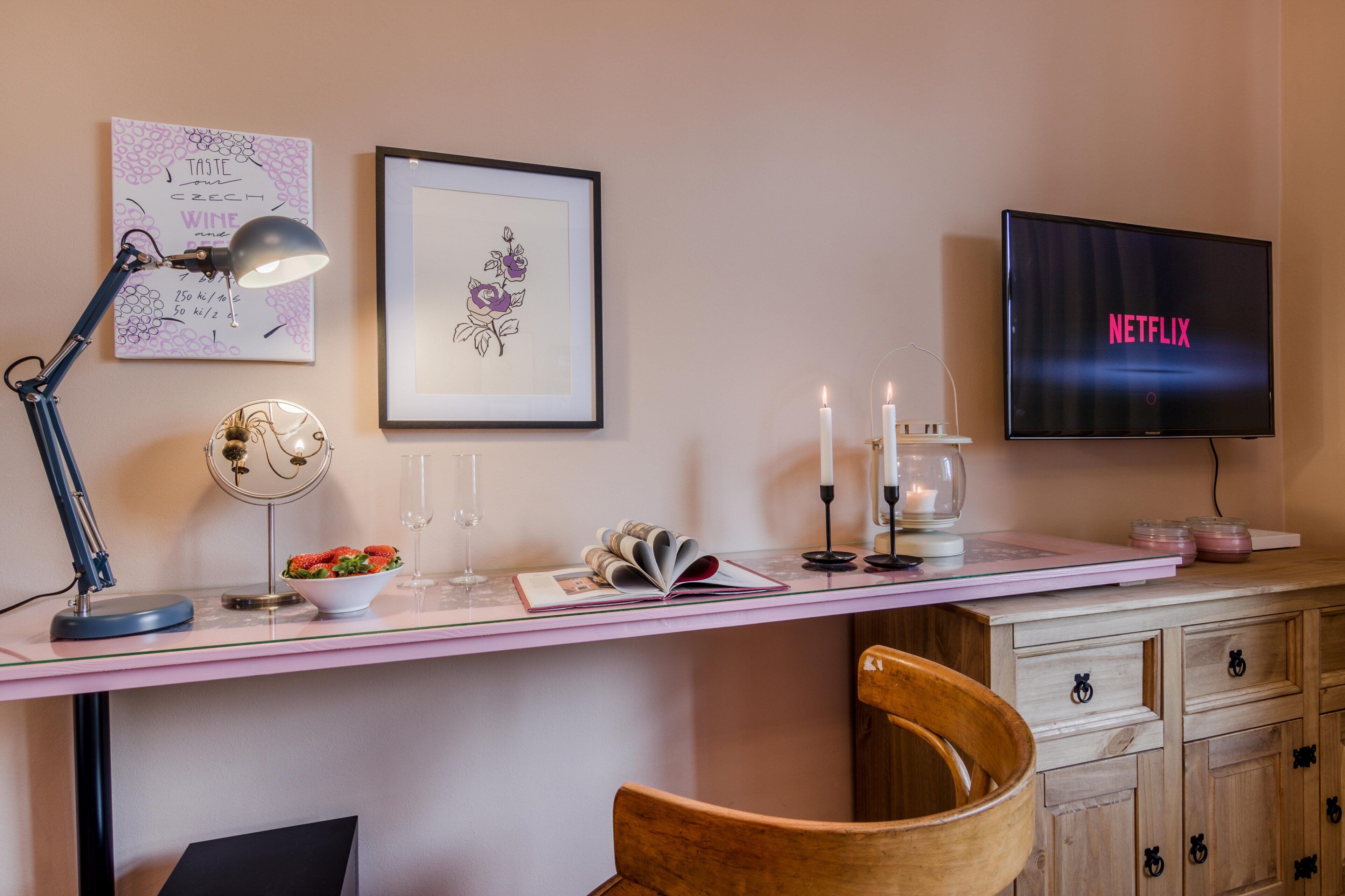 Una habitación moderna con escritorio, televisor mostrando 'NETFLIX', una obra de arte enmarcada, lámpara, velas y un bol de fresas.