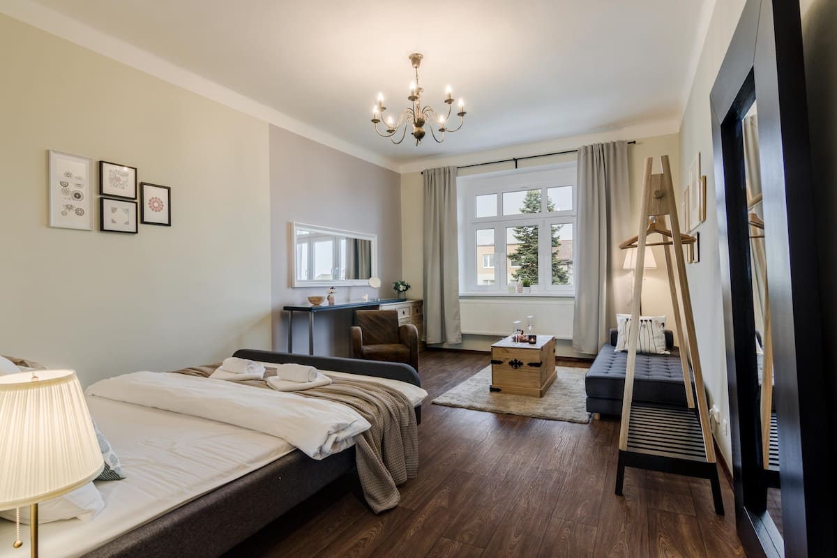Een moderne hotelkamer met een bed, nachtkastje, lamp en bureau met stoel. Er is een groot raam met gordijnen, een kroonluchter en kunst aan de muur.