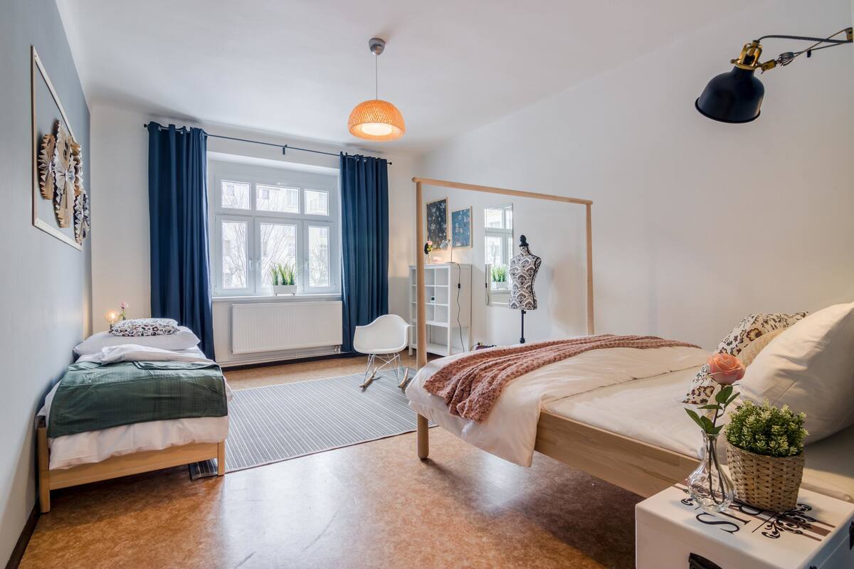 Een moderne slaapkamer met twee bedden, een groot raam met gordijnen, een hanglamp en een nachtkastje met een vaas en plant.