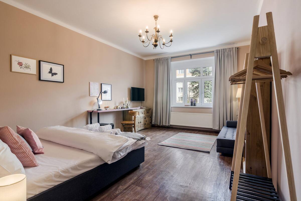 Een slaapkamer met een bed, nachtkastje, lamp, kapstok, raam met gordijnen en een houten schilderstandaard.