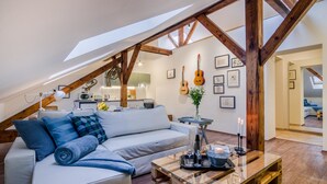 Ein gemütliches Loft mit geneigter Decke, Holzbalken, einem weißen Sofa, einem Glastisch und einer Küche im Hintergrund.