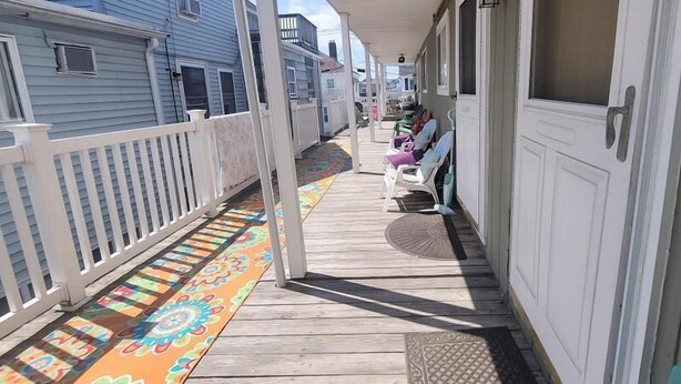 A covered porch with a colorful floor mat, white railings, and a row of white chairs with cushions. - Home Away From Home 2 (Hampton Beach)