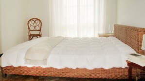 Un lit bien fait, recouvert d’une couette blanche, une chaise en osier et un canapé en osier dans une pièce dotée d’une grande fenêtre et de rideaux légers.