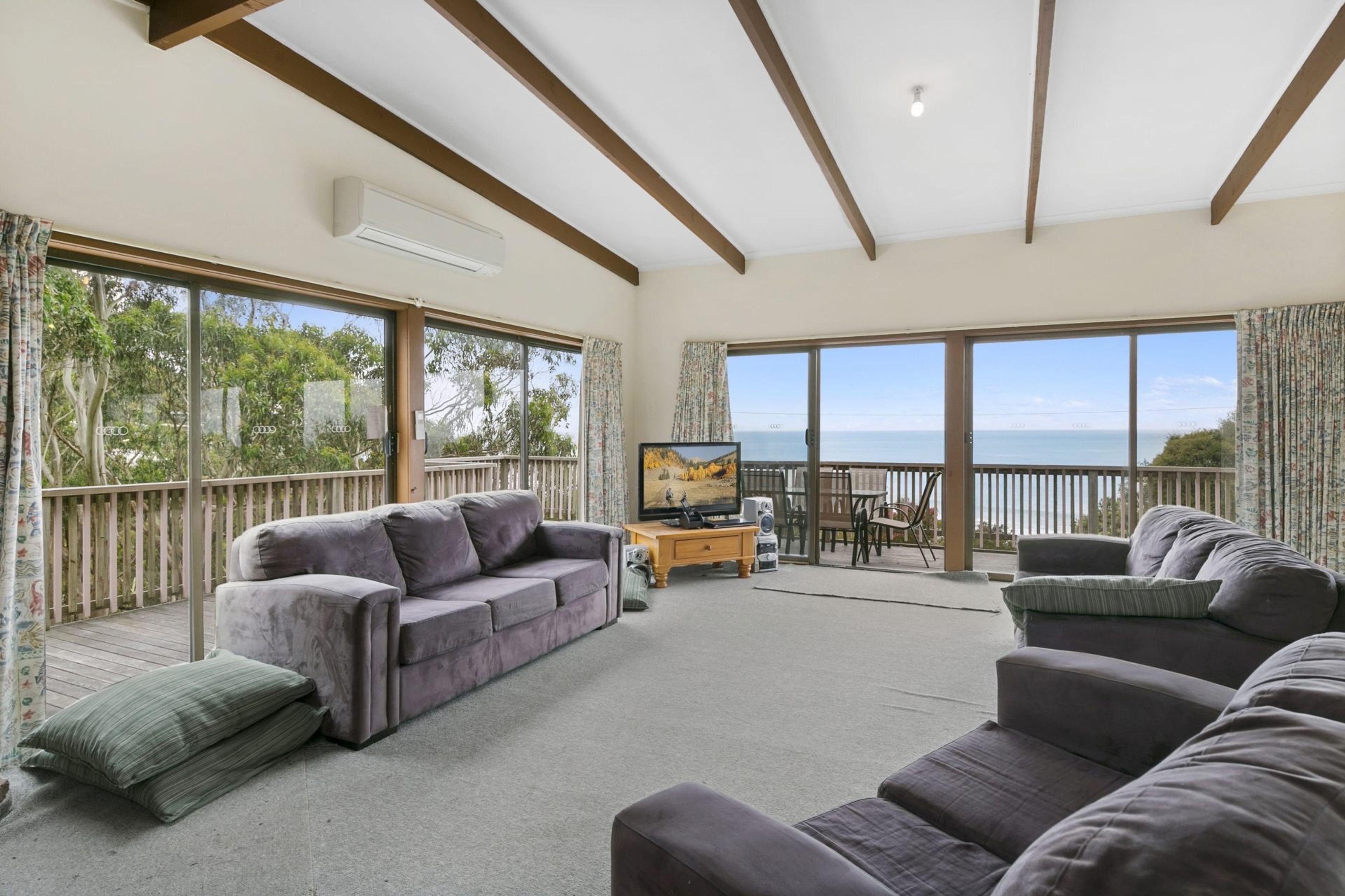 A spacious living room with a grey sofa, a television, and a view of the sea through large windows.