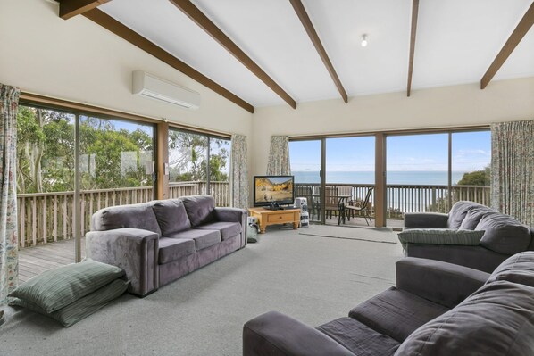 A spacious living room with a grey sofa, a television, and a view of the sea through large windows.