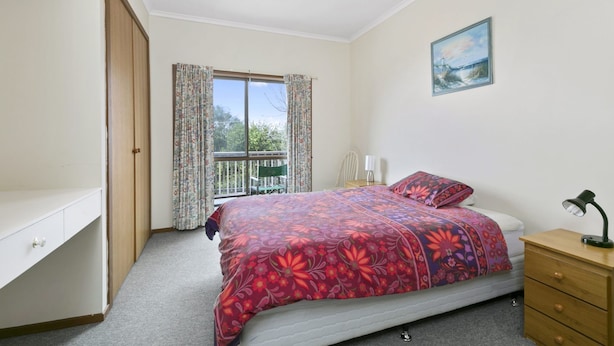 A bedroom with a bed, a desk, a chair, a window with curtains, a painting on the wall, and a balcony.