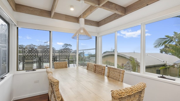 A dining area with a wooden table and woven chairs, large windows with a view of the outdoors, and a ceiling with exposed beams. - On The Deck (Lorne)
