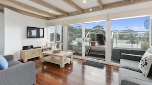 A spacious living area with a wooden floor, a sofa, a coffee table, and a television mounted on the wall. - On The Deck (Lorne)