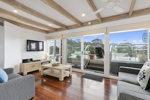 A spacious living area with a wooden floor, a sofa, a coffee table, and a television mounted on the wall. - On The Deck (Lorne)