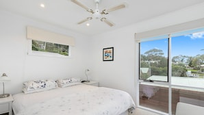 A bedroom with a large bed, a ceiling fan, a window with a view, and a bedside table with a lamp. - On The Deck (Lorne)