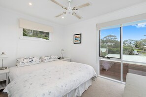 A bedroom with a large bed, a ceiling fan, a window with a view, and a bedside table with a lamp. - On The Deck (Lorne)