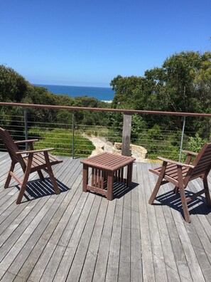 A wooden deck with two chairs and a small table, overlooking a coastal landscape with trees and a clear sky.