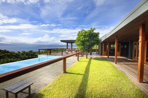 A modern outdoor pool area with a wooden deck, a grassy lawn, and a view of the ocean.