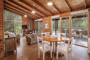 A wooden cabin with a dining area, a fireplace, and large windows.