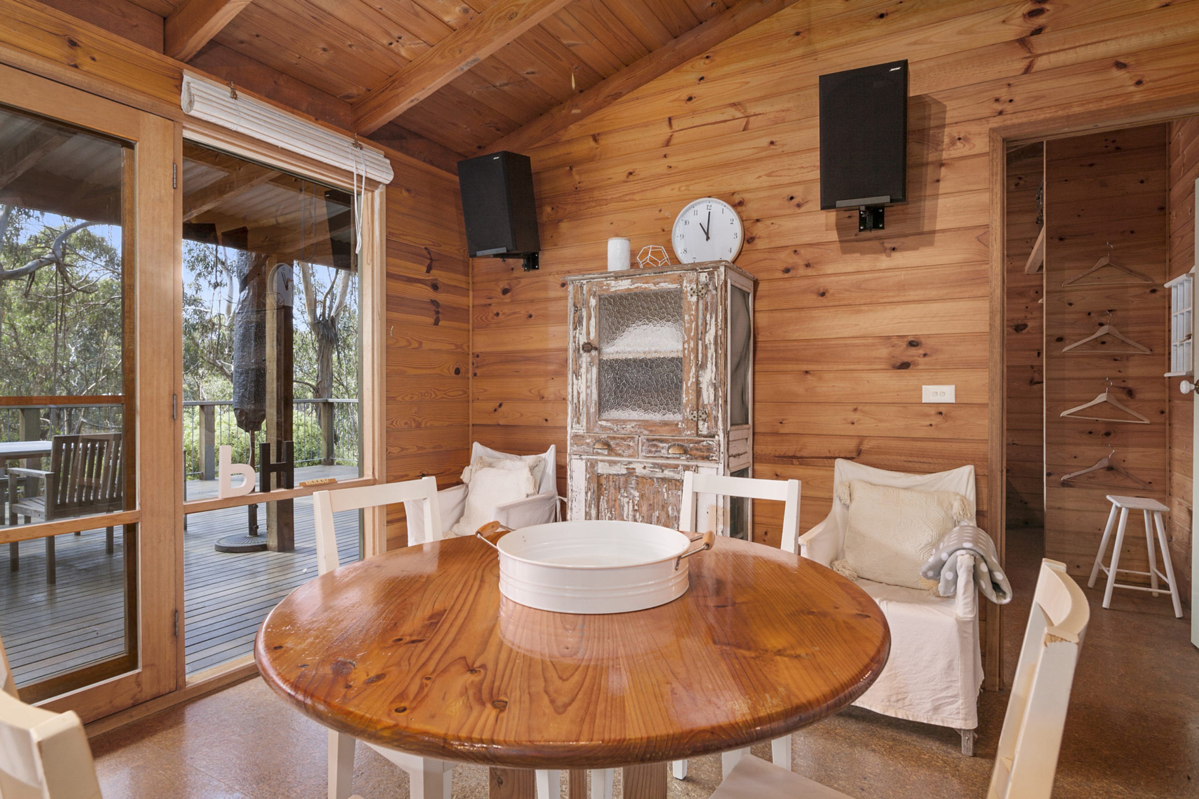 A wooden cabin interior with a round dining table, white chairs, a large window, and a clock on the wall.