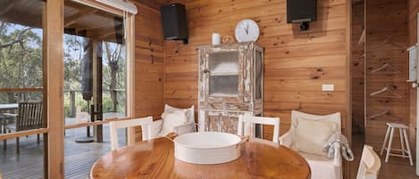 L'intérieur d'un chalet en bois, avec une table de salle à manger ronde, des chaises blanches, une grande fenêtre et une horloge au mur.