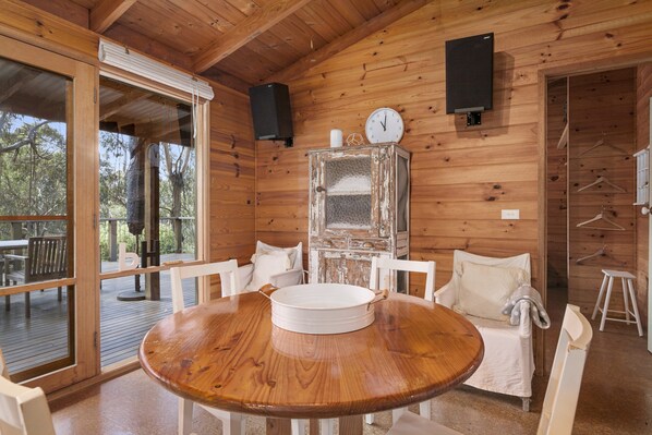 A wooden cabin interior with a round dining table, white chairs, a large window, and a clock on the wall.