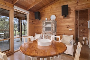 A wooden cabin interior with a round dining table, white chairs, a large window, and a clock on the wall.