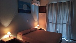 Un dormitorio con cama, dos lámparas de noche, mesita de noche, aire acondicionado, ventana con cortinas y un cuadro en la pared.
