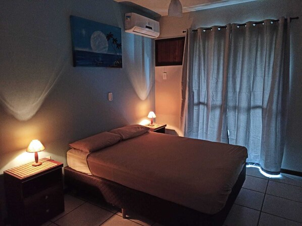 Ein Schlafzimmer mit einem Bett, zwei Nachttischlampen, einem Nachttisch, einer Klimaanlage, einem Fenster mit Vorhängen und einem Bild an der Wand.