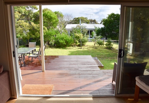 A sliding glass door leading to an outdoor deck with a table and chairs, surrounded by greenery and a house in the background.