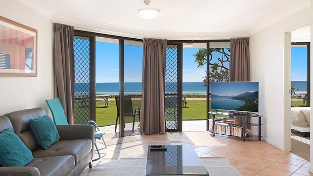 A living room with a sofa, a coffee table, and a TV. There is a sliding glass door leading to a balcony with a view of the sea. - San Simeon 6 - Ground Floor, Absolute Beachfront at Tugun! (Tugun)