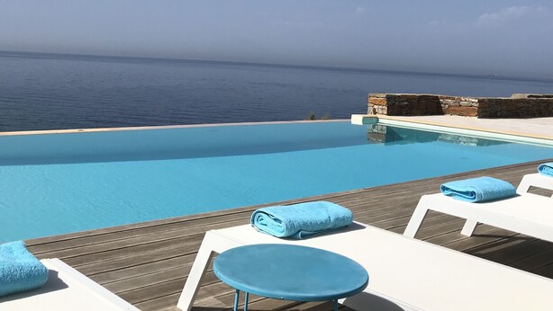 Une magnifique piscine à débordement d’un bleu profond, agrémentée de chaises longues blanches et de serviettes bleues, et offrant une vue imprenable sur la mer.