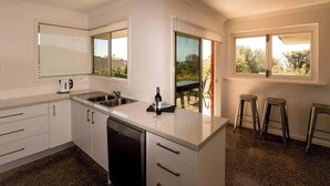 A modern kitchen with a dishwasher, a sink, and a wine glass on the counter. There are three stools near a sliding glass door leading to an outdoor area. - All Seasons—Coastal Charm Steps to Beach & Town (Dunsborough)