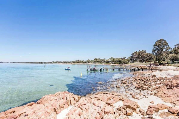 A rocky beach with clear water, a pier, and trees in the background.