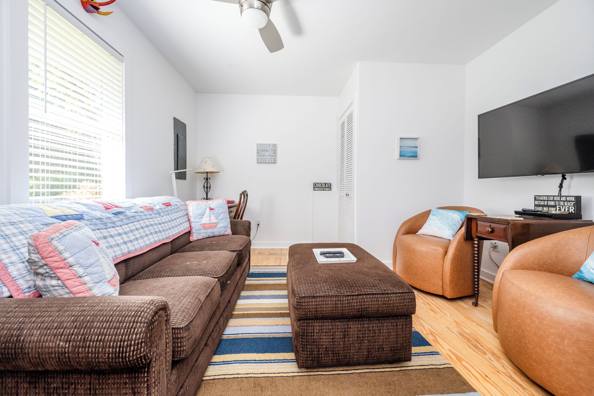 A modern, well-lit living room with a large window, a ceiling fan, a flat-screen TV, and a comfortable seating area with a sofa and ottoman.