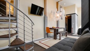 A modern living room with a grey sofa, a wooden staircase, a dining area with a table and chairs, and a flat-screen TV mounted on the wall.