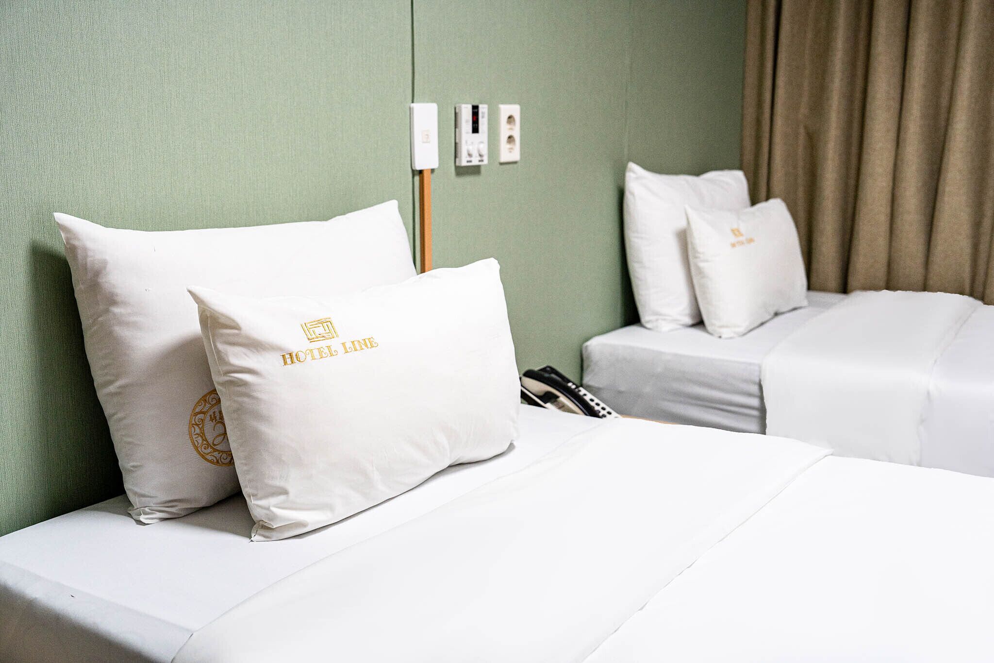 A hotel room with two beds, each with white bedding and matching pillows. There are electrical outlets on the wall and a curtain to the right.